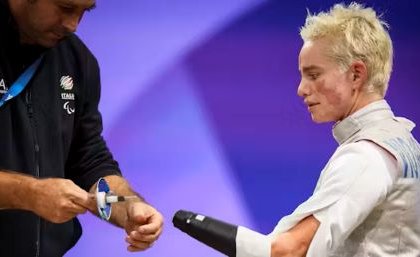 A female para athlete watches as her coach approaches with a prosthesis for her fencing competition. 